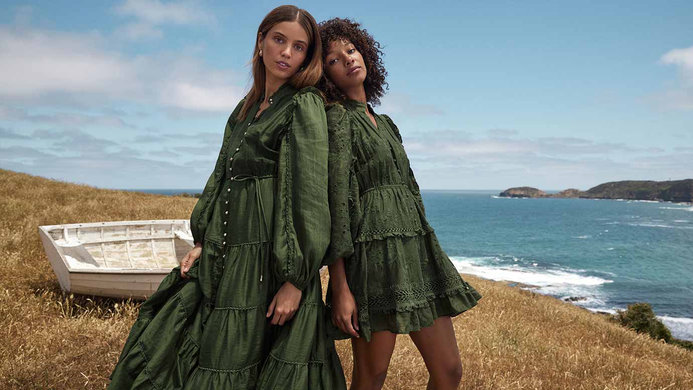 two models posing on a grassy cliff overlooking a turquoise blue ocean, a rusted whitewashed boat is in the background, they are wearing deep olive green long sleeve dresses, the black model has the mini length while the brunette model has a longer dress