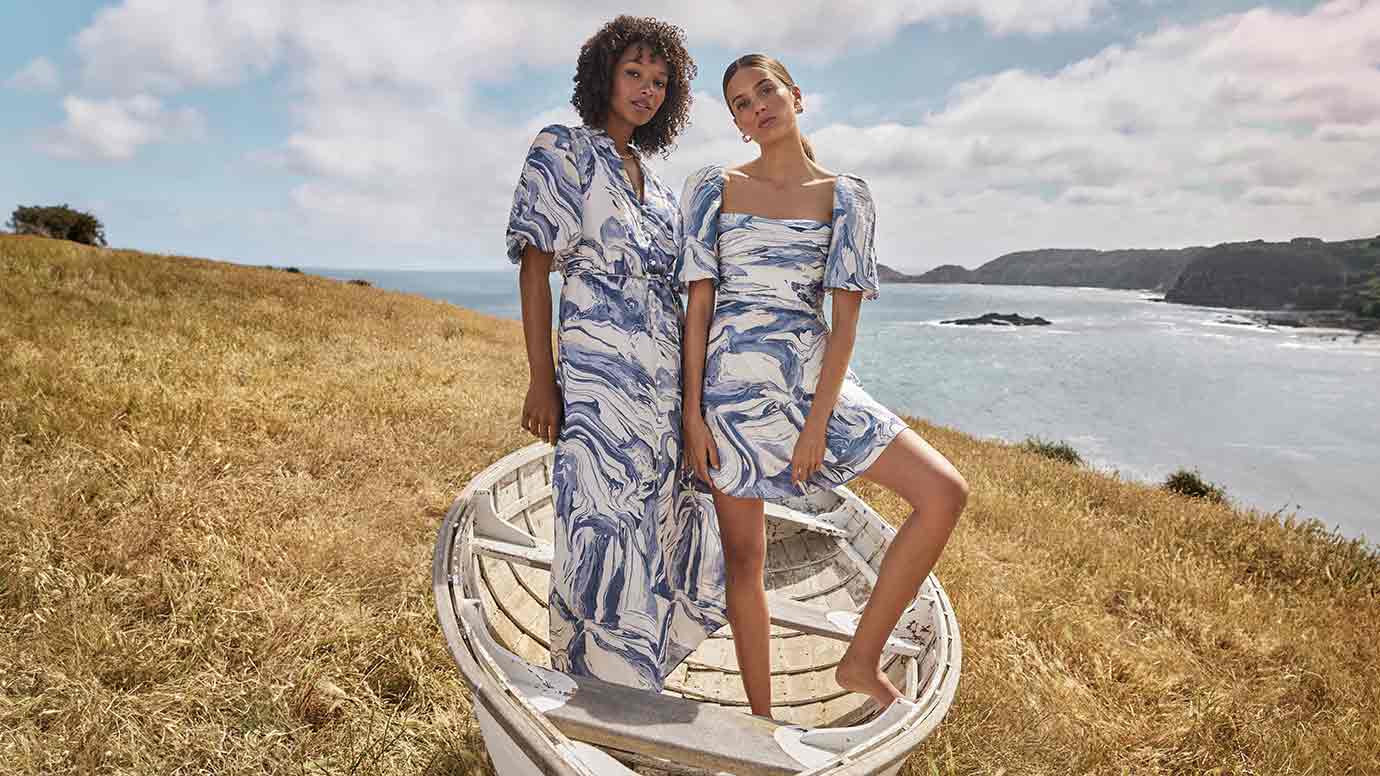 two models pose in a whitewashed old dingy style boat on a sun bleached grass-covered cliff overlooking a beautiful ocean, they both wear blue and white marbled print dresses, the black model has the longer maxi dress while the brunette wears the mini