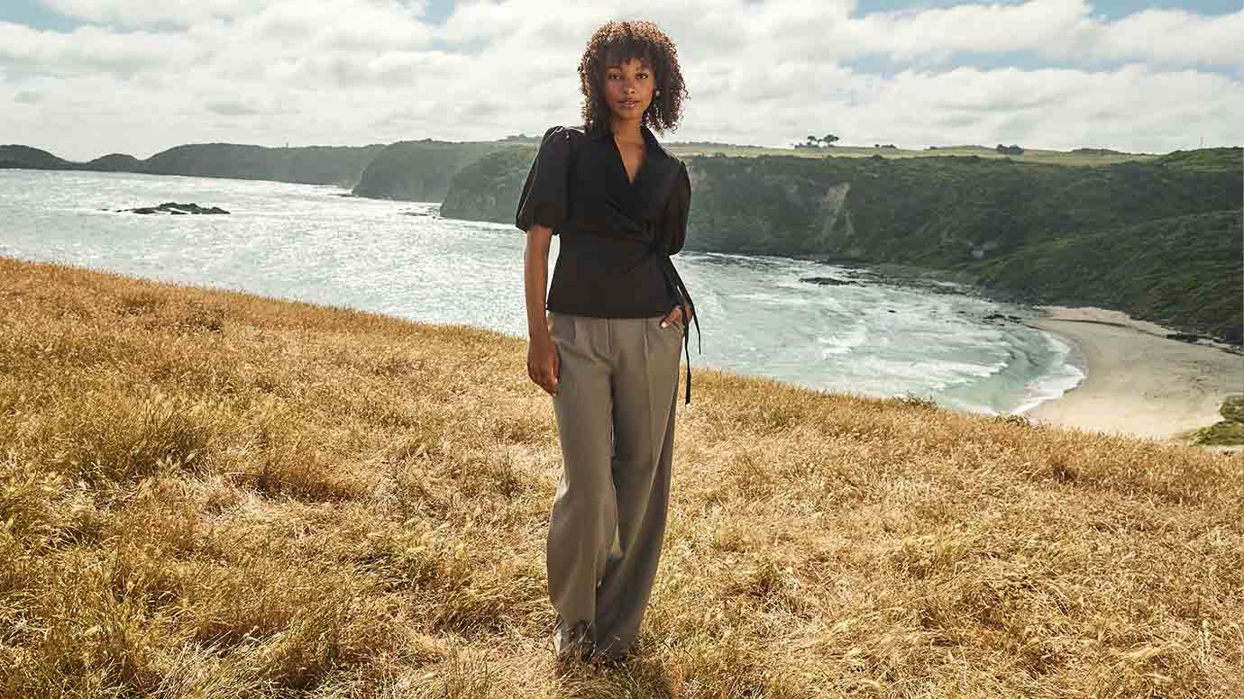 a model with soft curly afro hair poses on a sunlight cliff overlooking a beautiful ocean wearing a black wrap top and khaki/green wide leg pants
