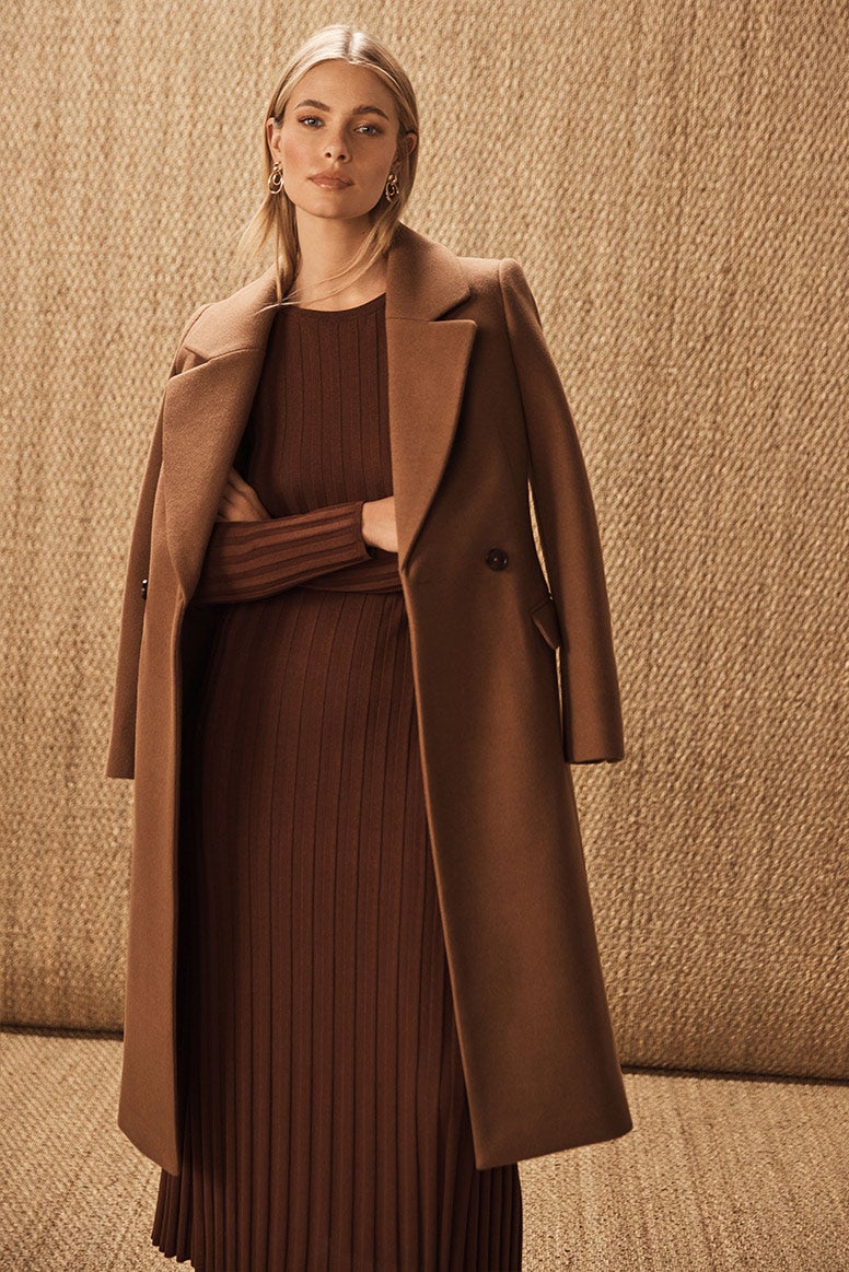 blonde model standing with arms crossed in a chocolate brown knit dress with wide ribbing and a chocolate brown coat draped on her shoulders, the background is a wall with jute fabric covering it