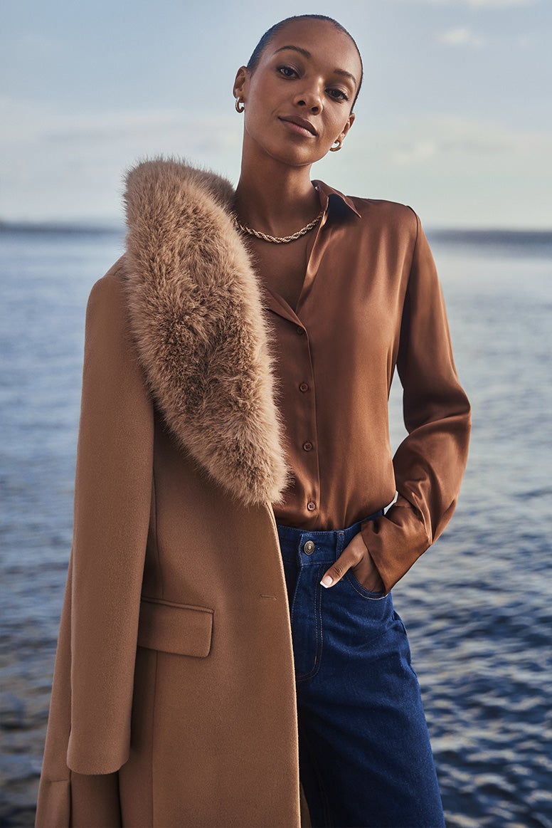 model in a caramel coat with faux fur collar with a brown satin shirt and blue jeans, blue sky and calm water is seen in the background