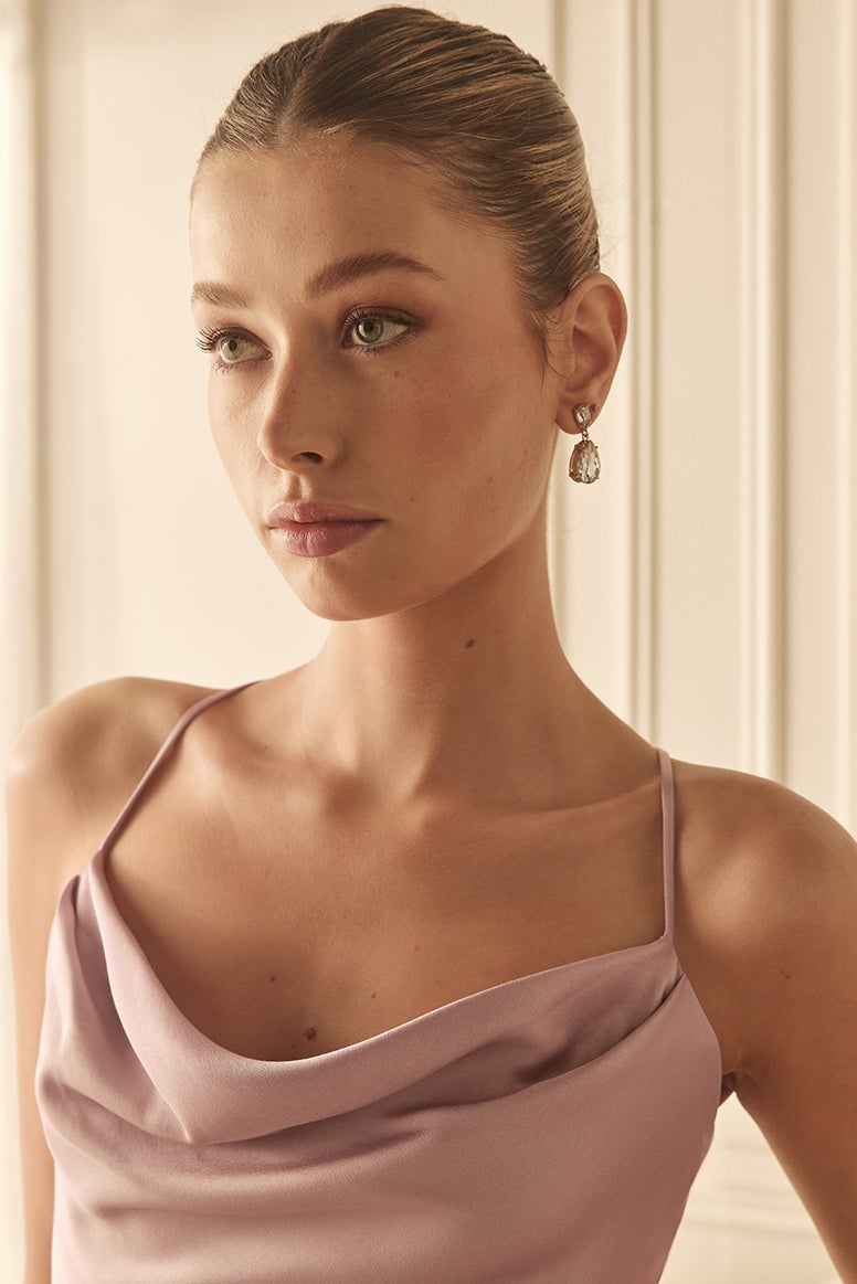 close up of a blonde models face, you can see she is wearing a strappy top or dress in soft pink satin with a draped cowl neck and drop earrings
