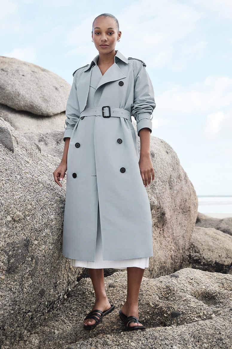 model with bronde hair wearing a light creamy natural crochet-esque beach dress but not transparent with a beige trench over the top, she is standing before a huge stack of boulders and rocks in an isolated coastal environment