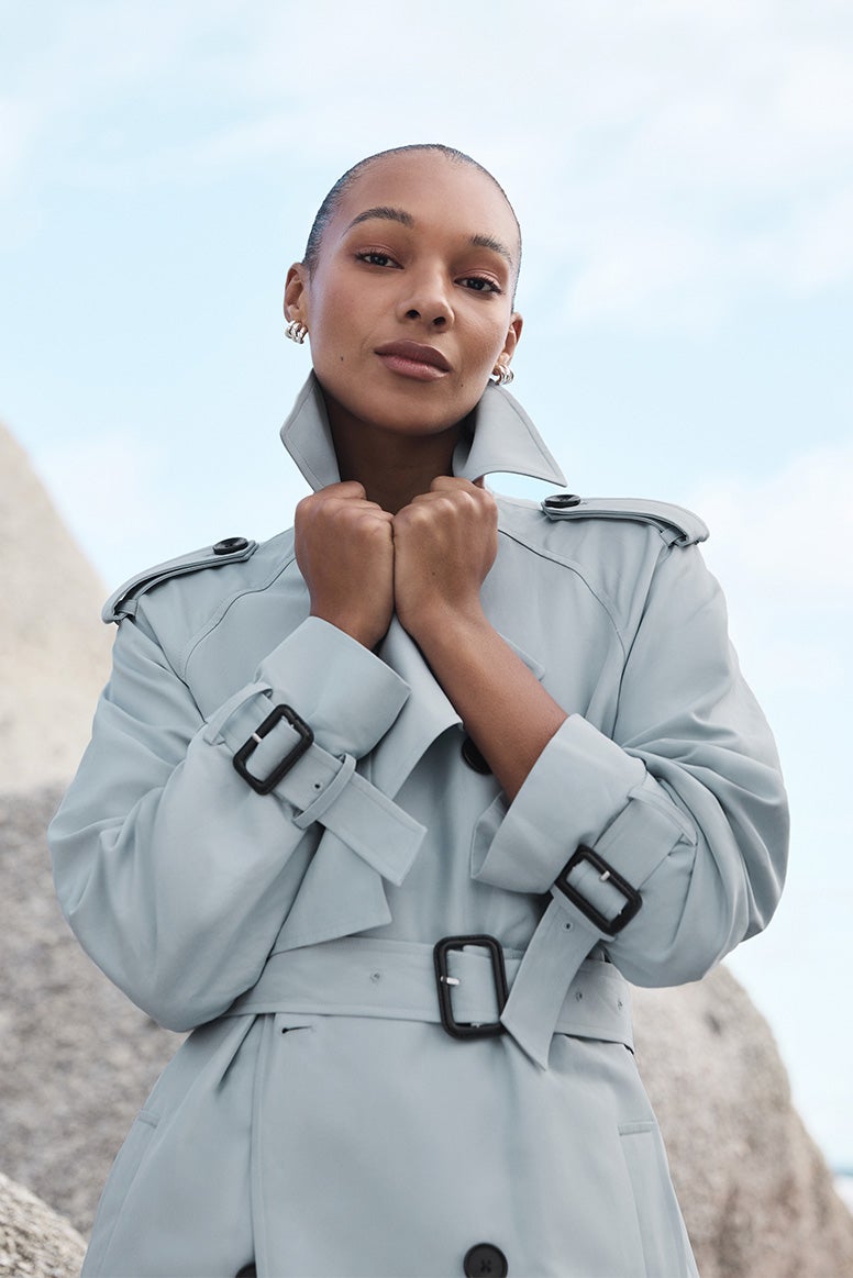 model with sandy coloured straight wearing a sand coloured trench, she is looking off into the distance and has her arms crossed
