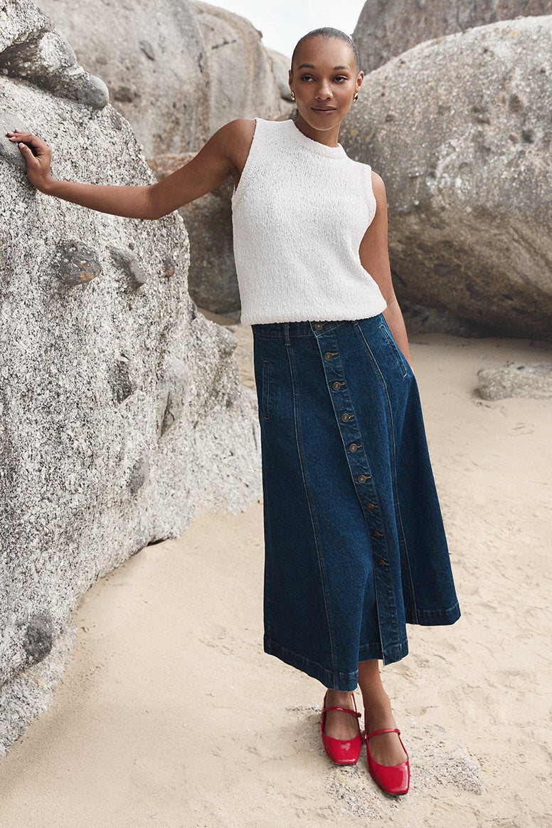 model with bronde straight hair wearing a light blue set that includes wide leg tailored pants and a longline strapless top with an asymmetrical hem, she is wearing white pointed heels, she is posing on sand in front of a large boulder