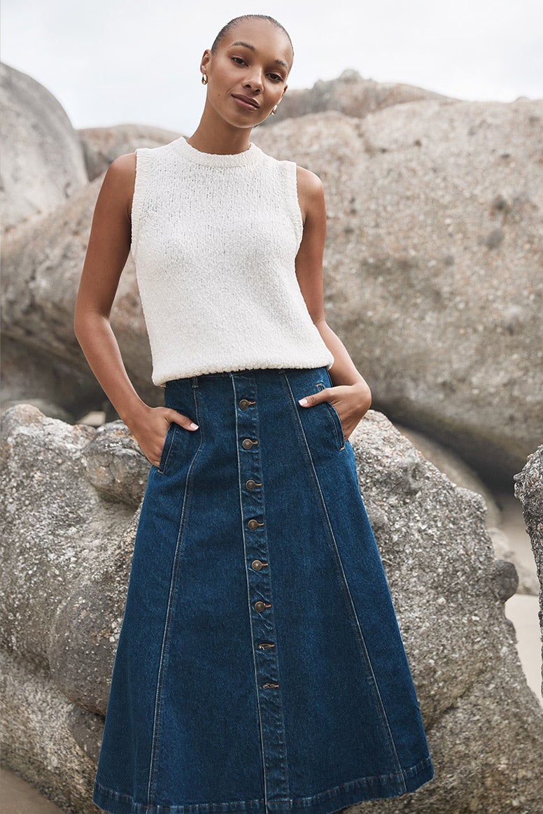 model with sandy blonde hair is standing before a large stone boulder, she is wearing light blue tailored pants and a matching strapless top and a cream trench