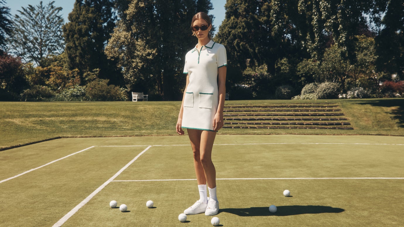 model is standing in profile on a lush green tennis court, she wears a white knit dress with green contrast tipping and sunglasses, white socks and white sneakers, white tennis balls are all around her feet
