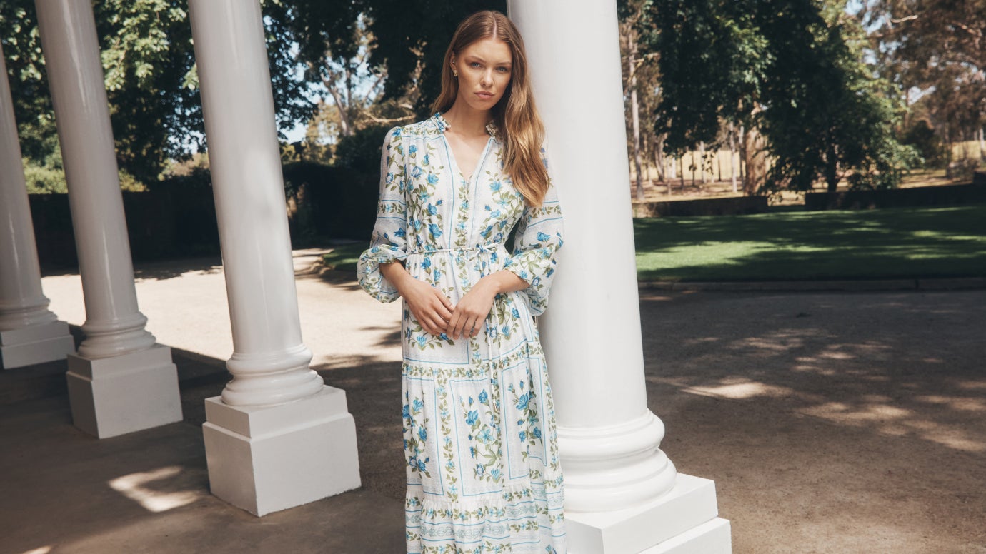 model wear long sleeve flowing white dress with delicate placement print florals in blue and green, she leans against grand white columns