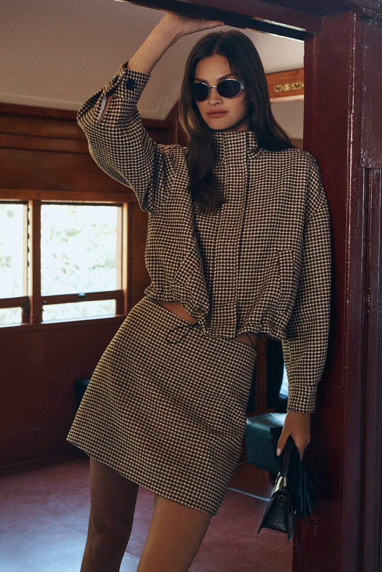 Model is standing inside the train carriage in a matching check set, holding a mini bag in her hand as she leans against the door frame in a pair of dark sunglasses.