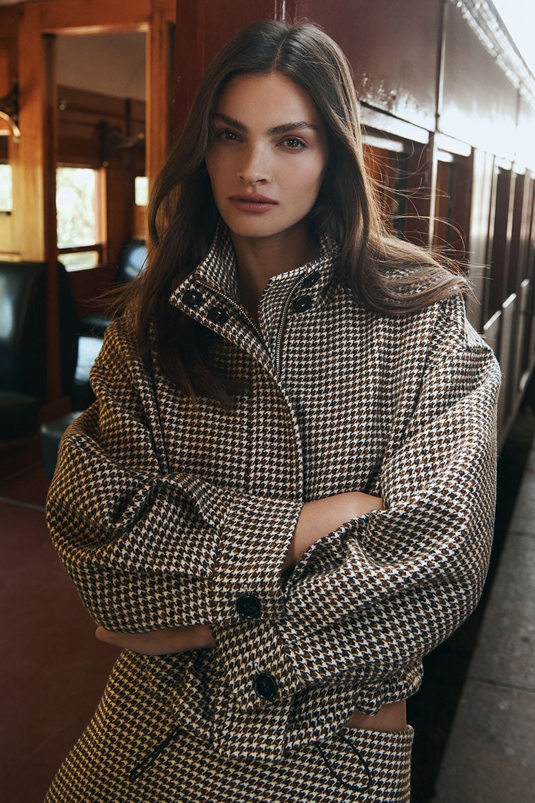Model stands with arms crossed wearing a matching check set. She leans against the doorframe of the red, vintage train carriage looking into the camera.