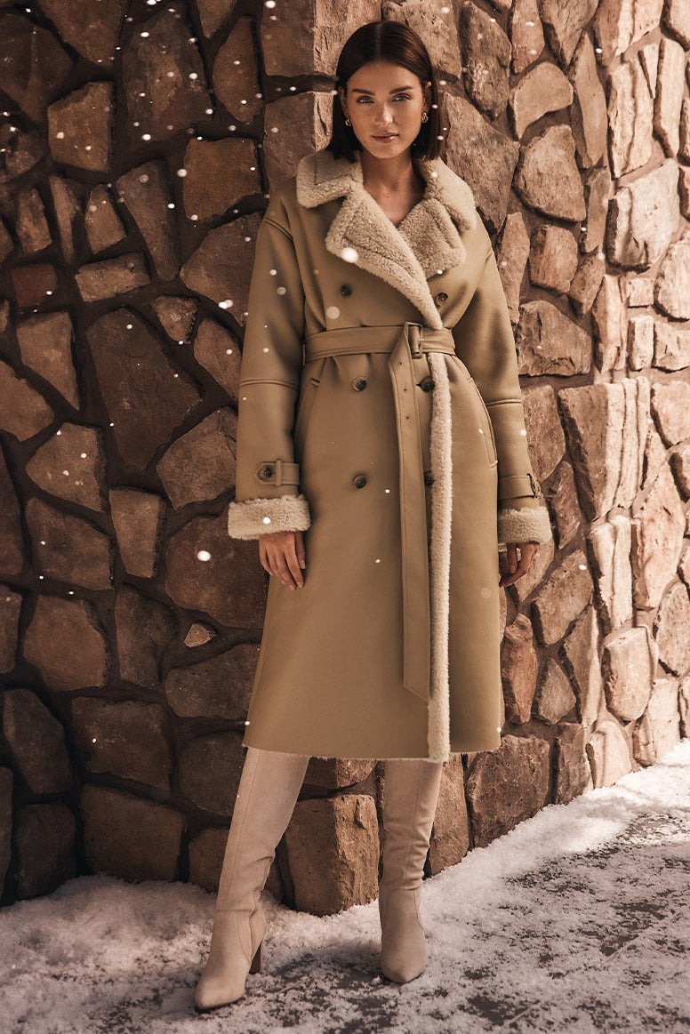model in a longline camel coat with shearling trims and knee high camel boots standing against a stone wall with snowflakes gracefully falling over her on the left side of the photo