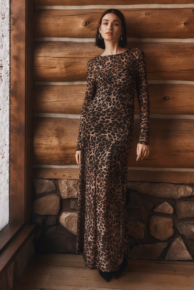 model with brown bob standing against a log cabin wall in a long sleeve maxi dress in leopard print, snow can be seen on the window to the left