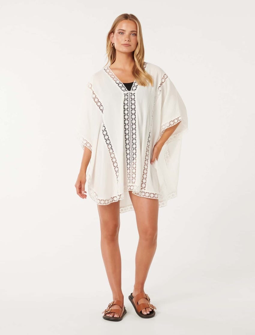 a model with blonde hair poses in a white beach cover up poncho with cut out embroidery detailing and brown slides with black soles and large buckles