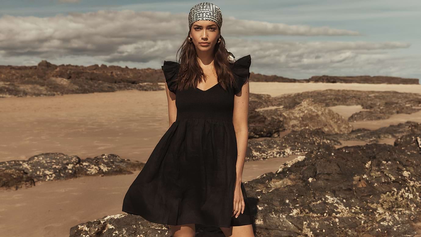 model with long dark hair wearing a short black dress with ruffled shoulders, she poses in front of rocks, sand and the ocean and wears black and white silky headscarf