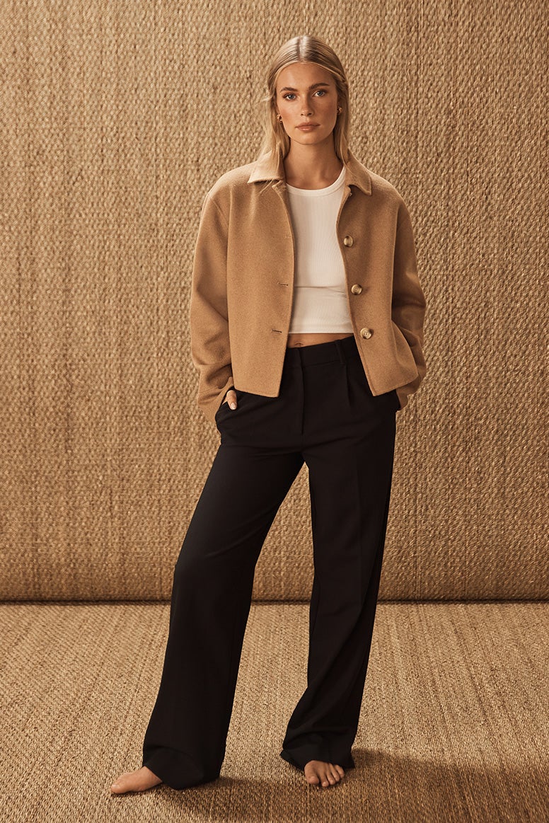 model standing barefoot posing wearing black pants, white basic top, and a crop camel jacket, the background is a photography studio with natural jute coverings on the floor and wall