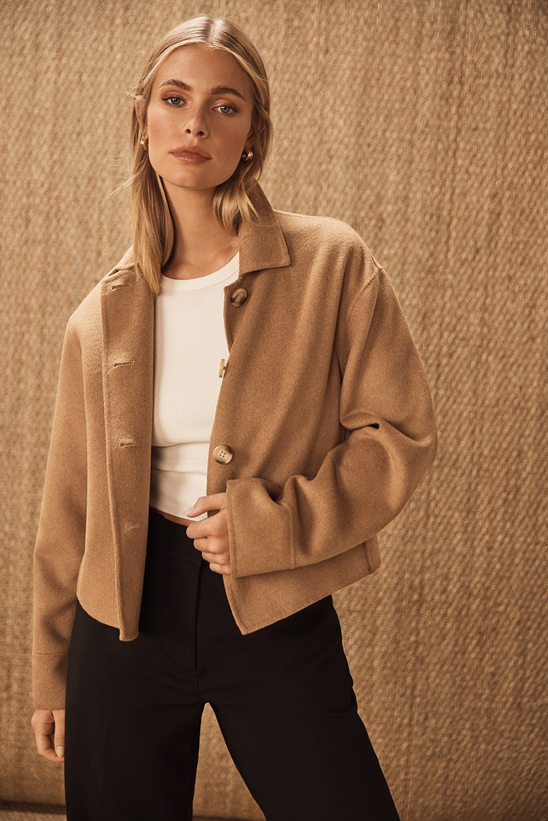 model posing wearing a in a white basic top, camel cropped jacket and black tailored pants, the background is photography studio with natural jute coverings on the floor and wall