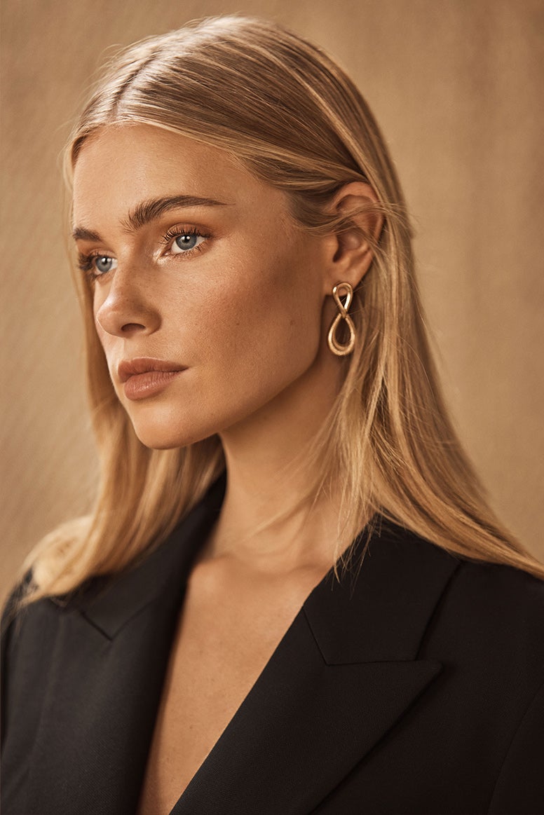 close up of models face she is wearing a gold infinity loop style earring with her hair tucked behind her ear, you can just see the top of her black blazer jacket