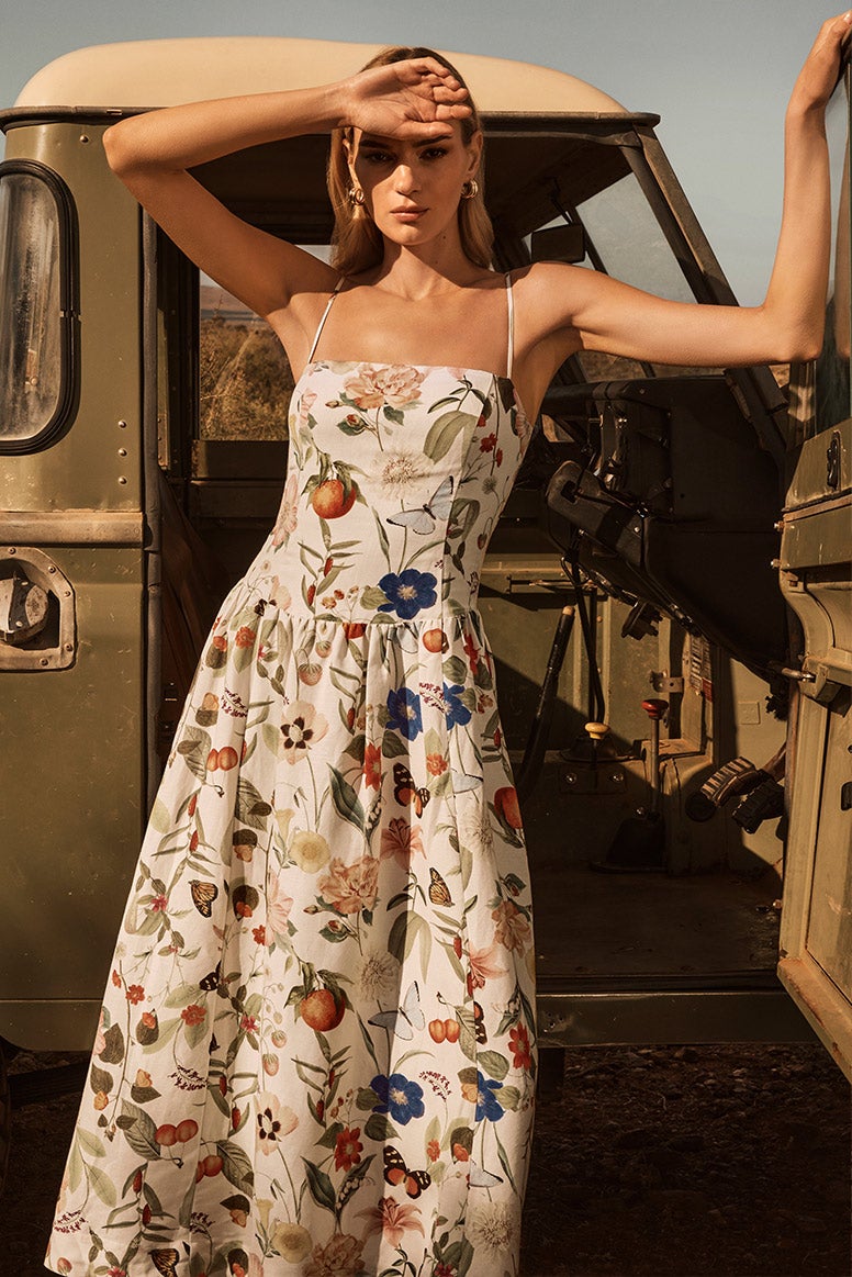 model with wearing long strappy dress with a multi colour floral print shades her eyes from the sun, she is leaning on the open door of a vintage army green pickup truck