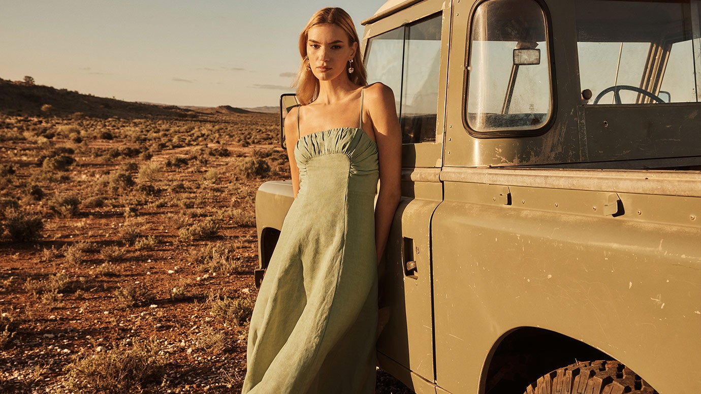 model poses with hands behind her back leaning against the side of a army green vintage pickup truck in a dusty landscape, she is wearing a pistachio green linen dress with thin straps and gathering along the bust 