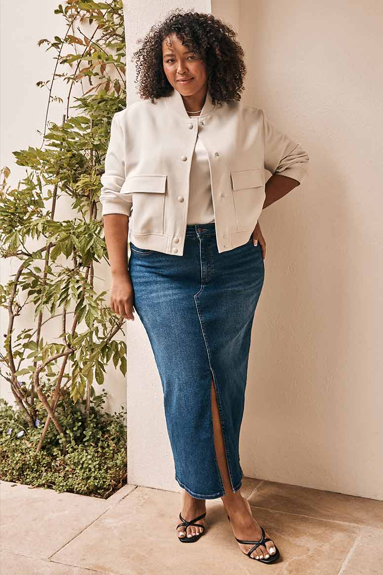 curve model with curly textured hair wearing a cropped cream bomber jacket, cream top, and a long denim skirt with black flip flop style mule heels, she poses in front of a beige wall with greenery in the background