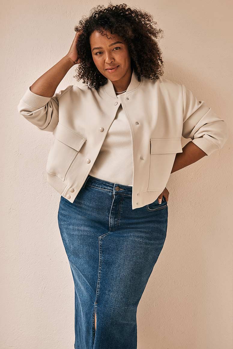 curve model with curly textured hair wearing a cropped cream bomber jacket and a long denim skirt posing with one hand to her head in front of a beige wall 