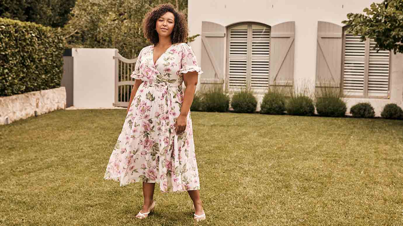 plus size model with curly textured hair wearing a white wrap dress with green and pink floral and white heels, she is standing on a manicured lawn with a large home behind her