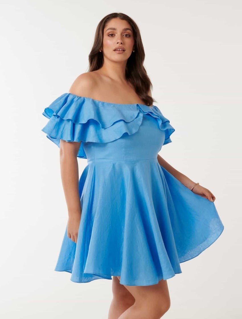 plus size model with long brunette hair poses against a white backdrop in a blue linen mini dress with an off the shoulder ruffle design