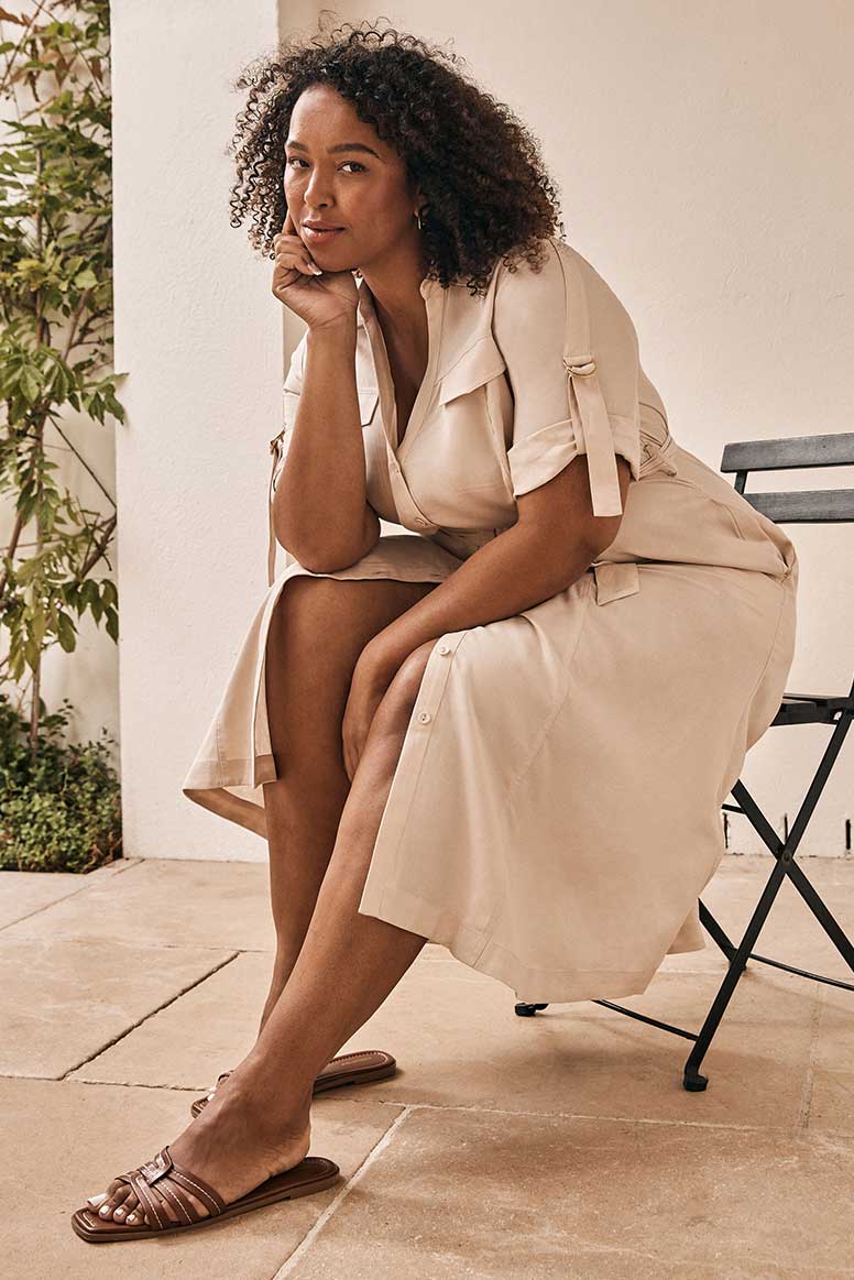 curve model with curly textured hair wearing a camel coloured shirt dress with utility inspired detailing sitting on a wooden and metal patio chair, a beige wall and greenery is seen in the background 