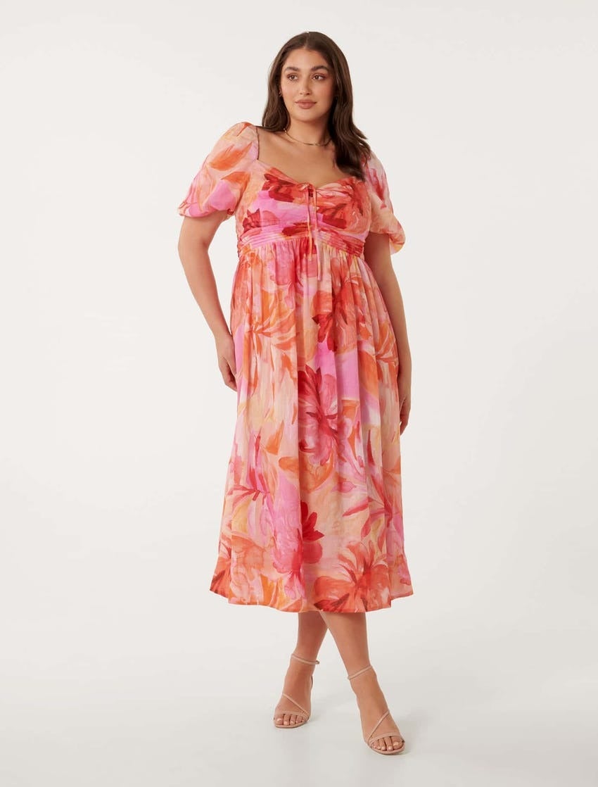 a plus size brunette model poses against a white backdrop in a pink and peach puff-sleeve dress with a large scale tropical floral print 