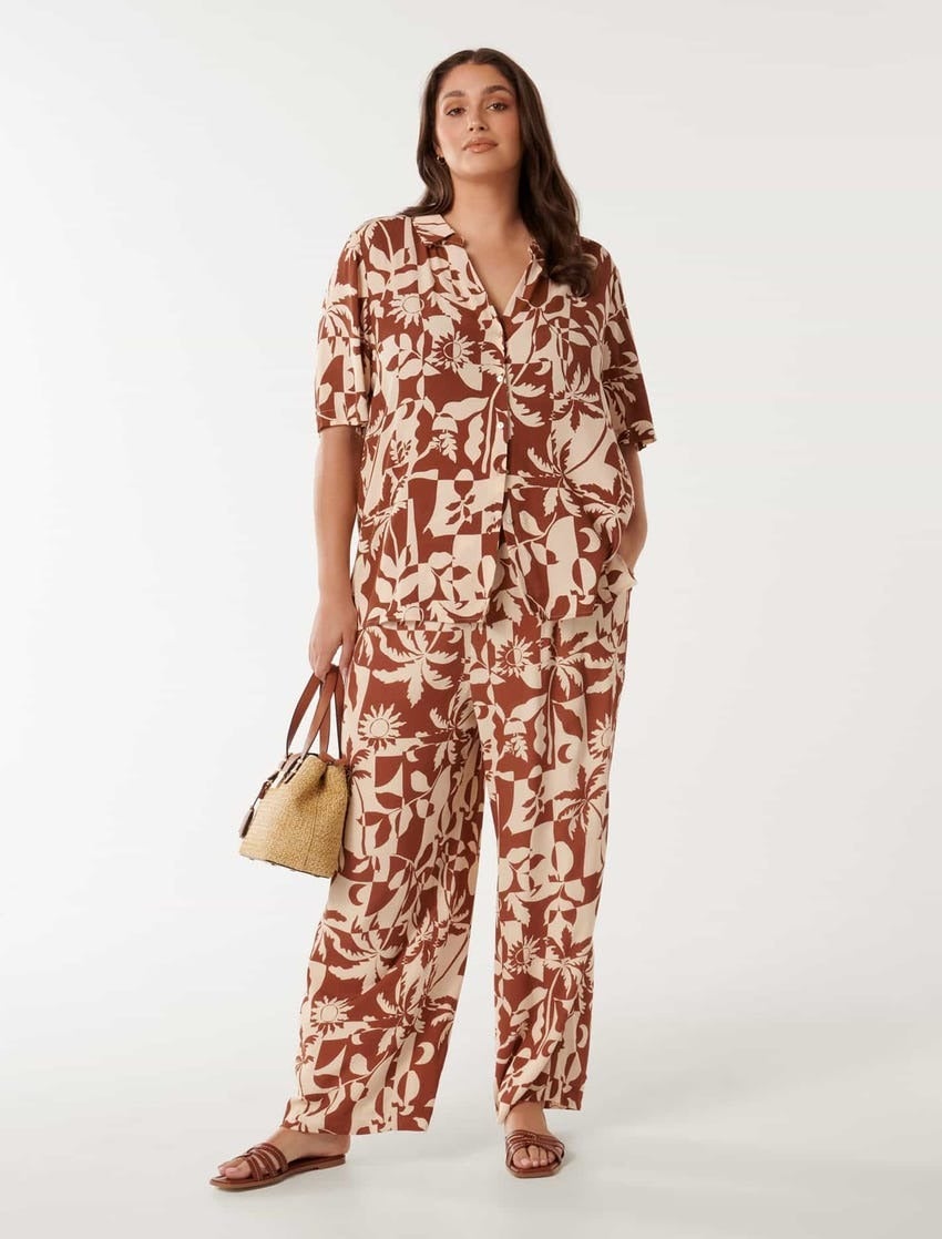 plus size model poses against a white backdrop in a brown and white resort set consisting of a button down shirt and loose pants, the matching print is an abstract one that features palm, sun, and moon motifs 