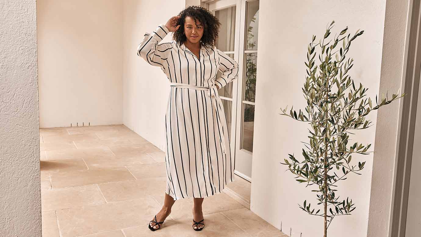 curve model with curly textured hair wearing a white satin shirt dress with think black vertical lines and black slip on strappy heels, she is posing in a beige patio space with a large plant to the right of her