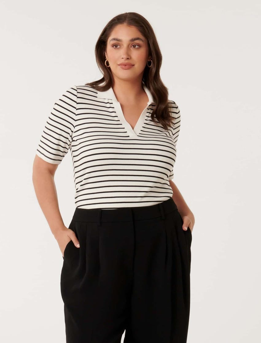 a brunette plus size model poses against a white backdrop wearing white and black striped polo with a white collar tucked into black pants