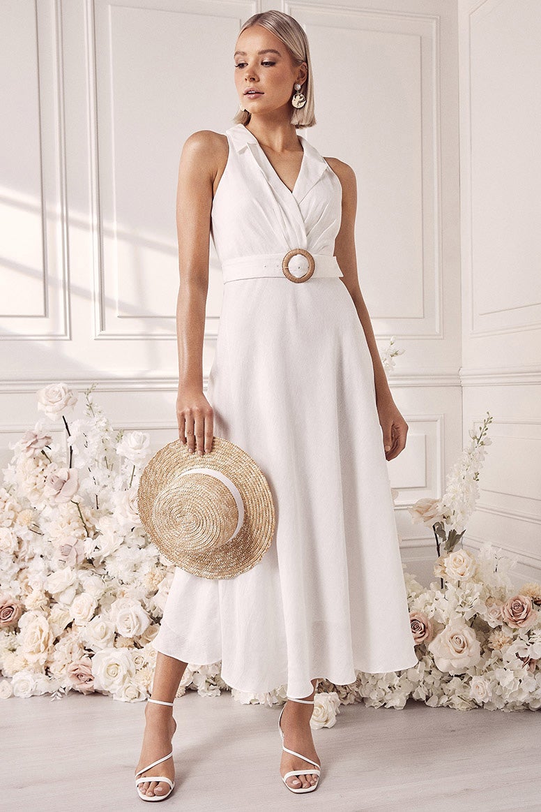 a model with a blonde bob wearing a white sleeveless linen dress with a collar and a belted waist poses in front of a white floral installation and panelled walls, she also wears strappy white heels and holds a straw hat