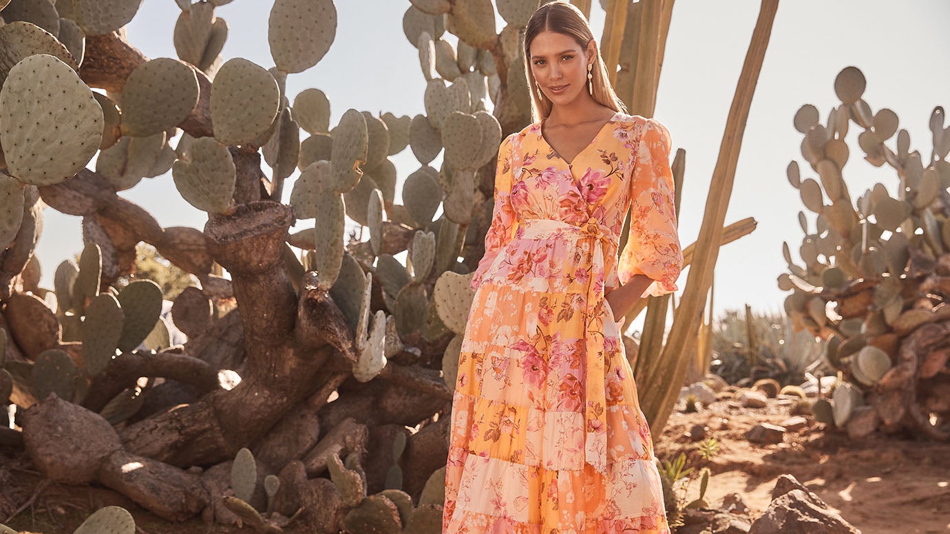 model with long hair poses in front of a large cactus with circular petals in a desert landscape wearing a floaty long sleeve dress in tiled floral print in citrus and pink shades 