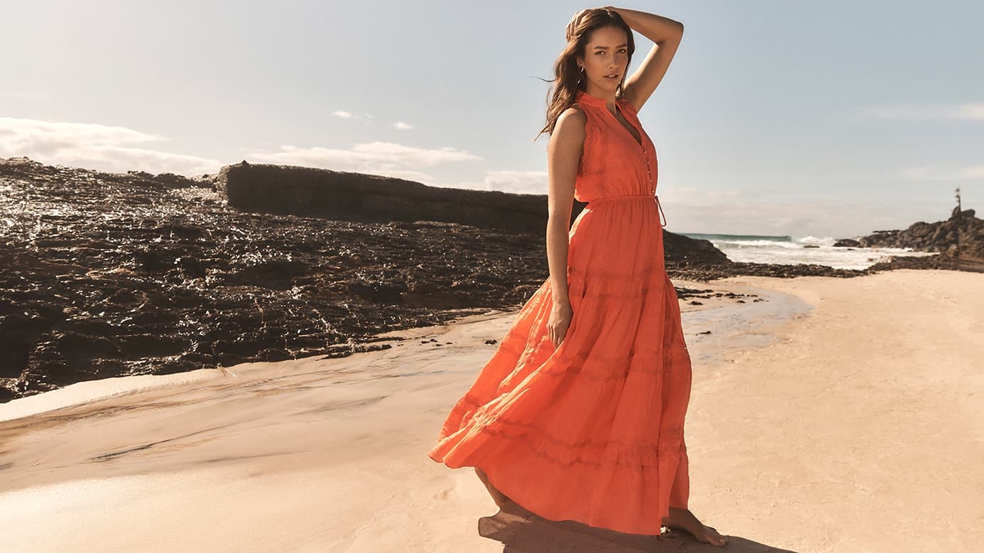 model with long hair wearing a long tiered orange maxi dress stands barefoot posing on golden sand with the water and black rocks behind her