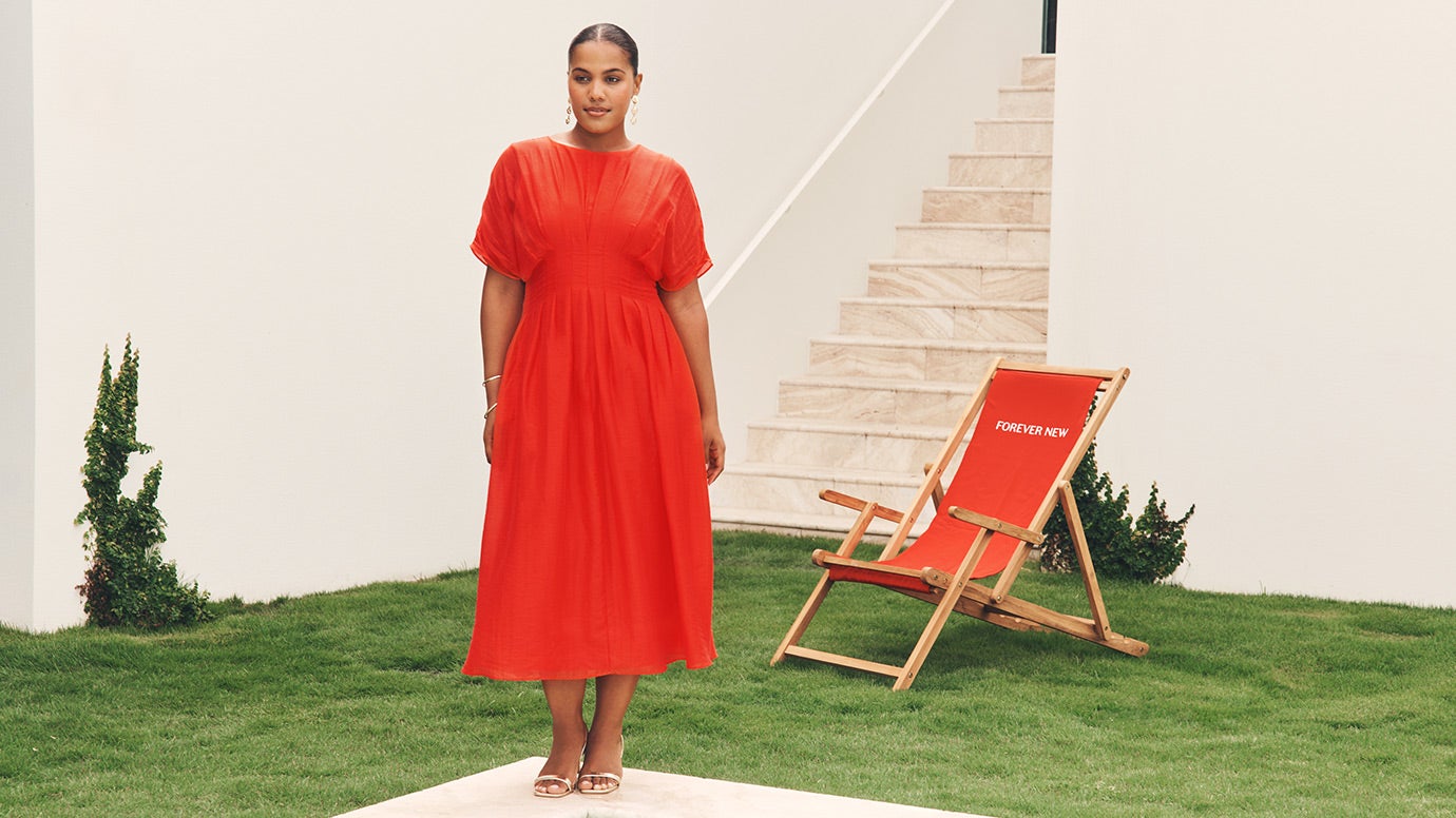influencer and model sierra johnson wearing a flowing red dress with draped kimono style short sleeves and a seamed waist to give a cinched look, she is standing near a manaicured lawn and a red sunlounger