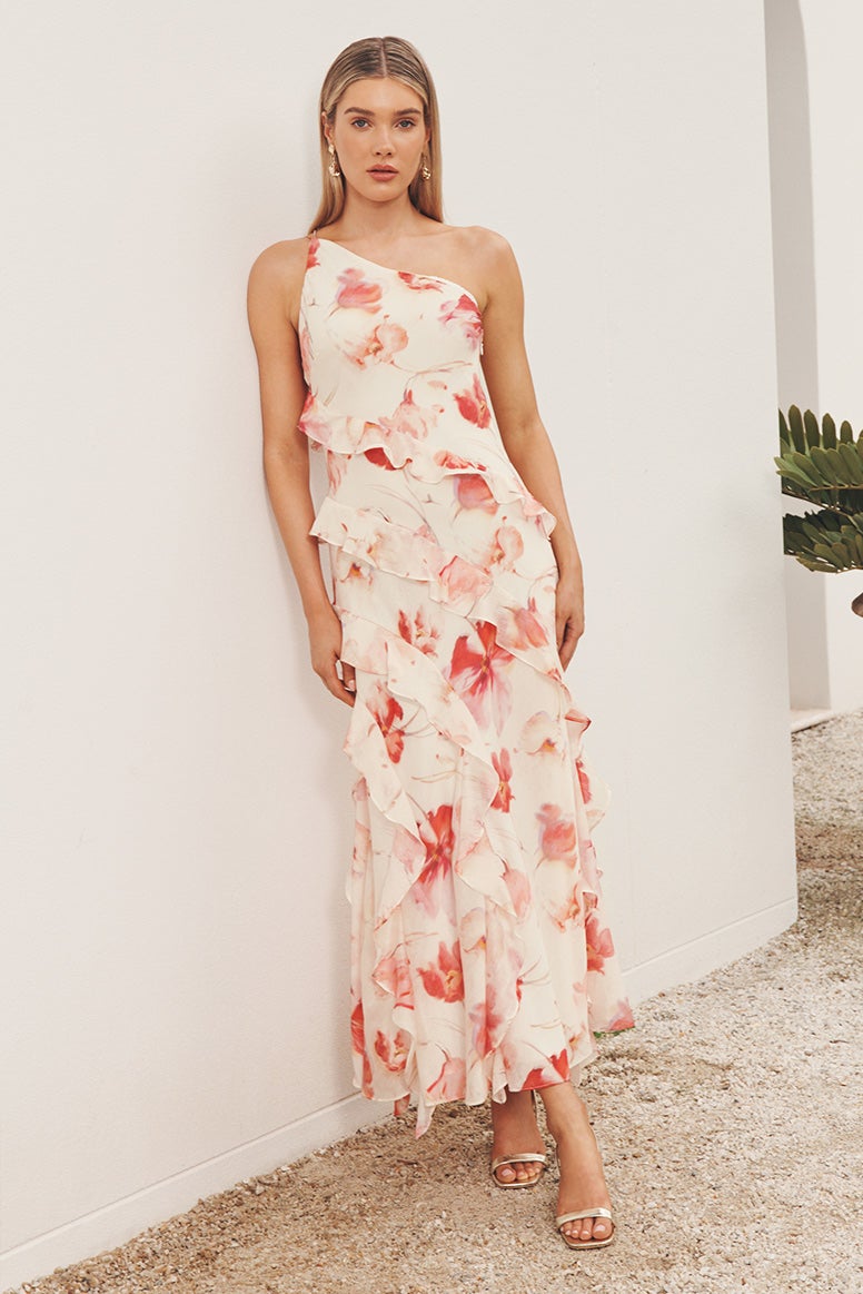 influencer and model ellie faust wears a one shoulder ruffle maxi dress in a floral print, she has long blond hair and is standing on cream pebbles in front of a white building