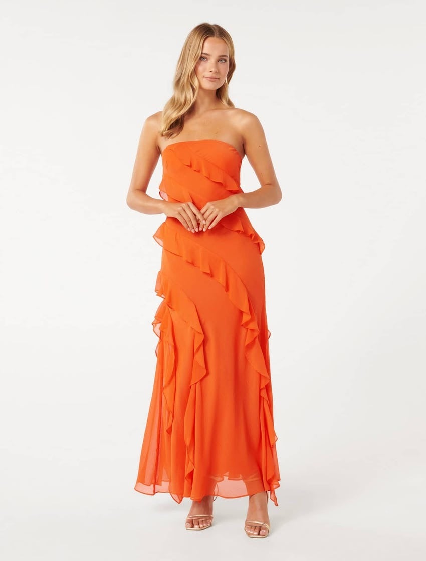 blonde model poses against a white backdrop clasping her hands with her elbows out wearing a long bright orange strapless event dress with soft drapey ruffles