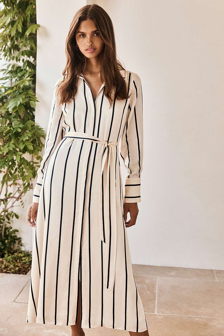 model with long dark hair wears a satin shirt dress with vertical black and white stripes and a sash belt 