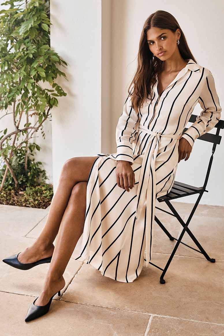 model with long dark hair sitting on a patio chair wearing a cream satin shirt dress with thin black stripes that run vertically and pointed toe black kitten heels