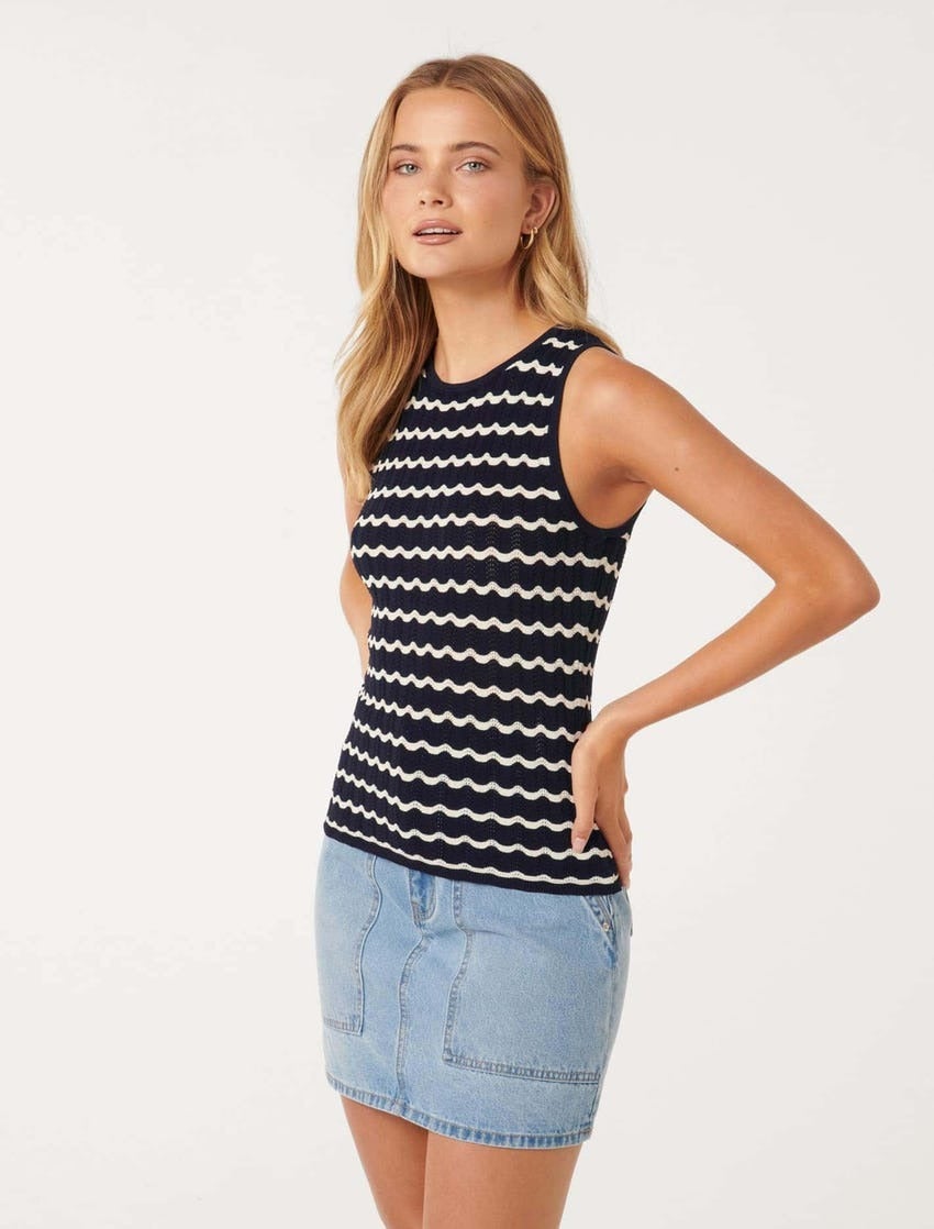 a blonde model poses with hands on hips against a white backdrop wearing a navy tank top with wavy white lines across it, she is also wearing a light wash blue denim mini